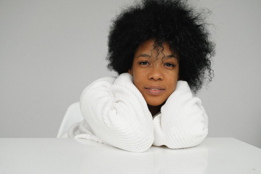 Charming African American female in white sweater sitting at table and cuddling while looking at camera on on gray background