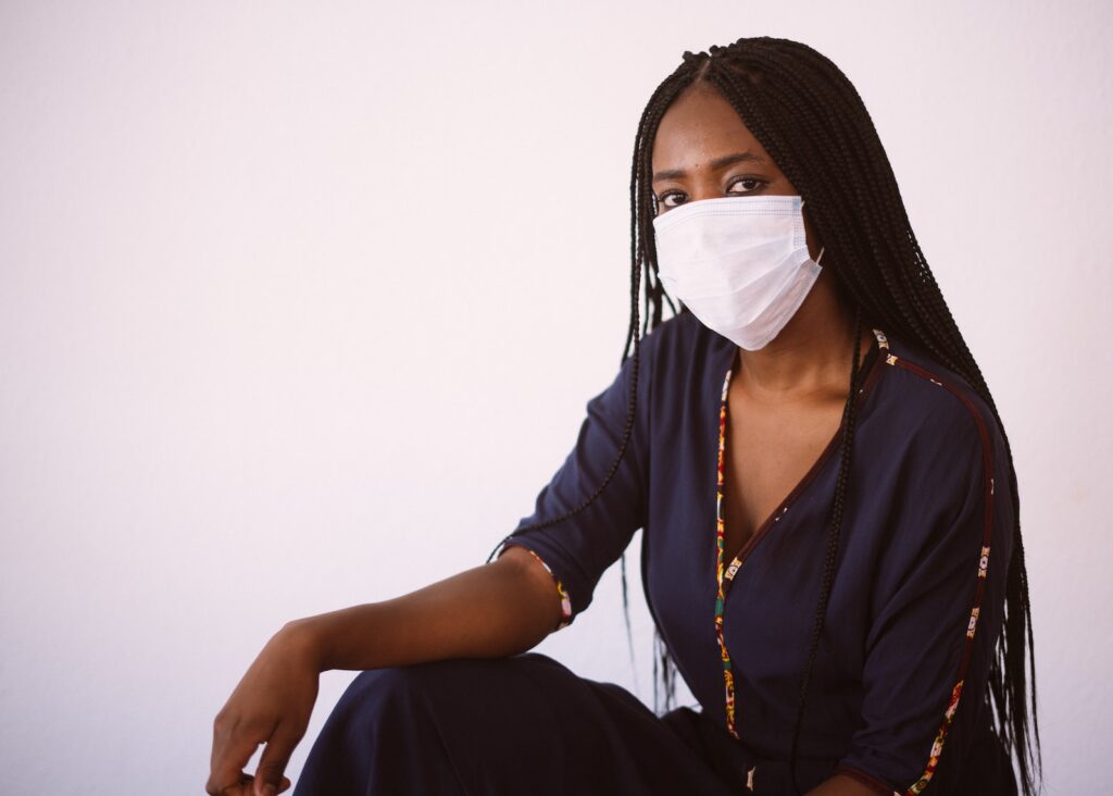 Side view of unrecognizable African American woman in casual dress and protective mask sitting against gray background looking at camera