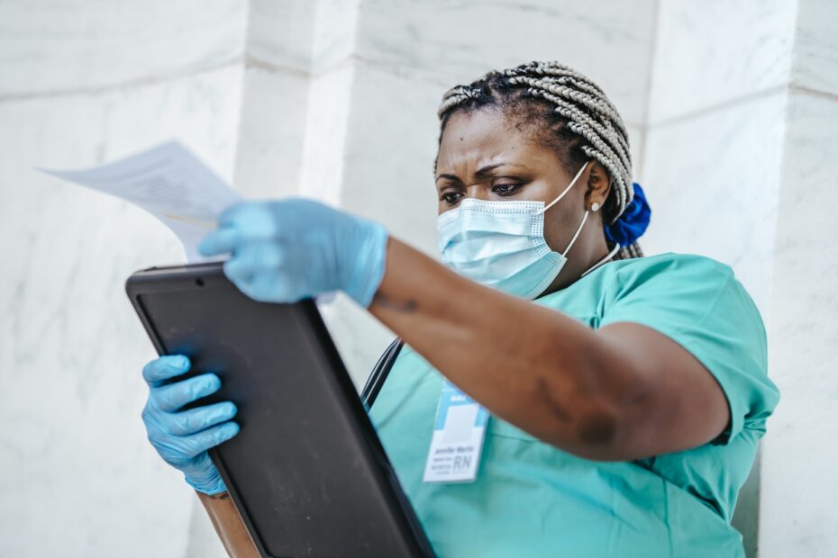 Low angle of focused black female nurse in mask and gloves standing in hospital and looking through papers
