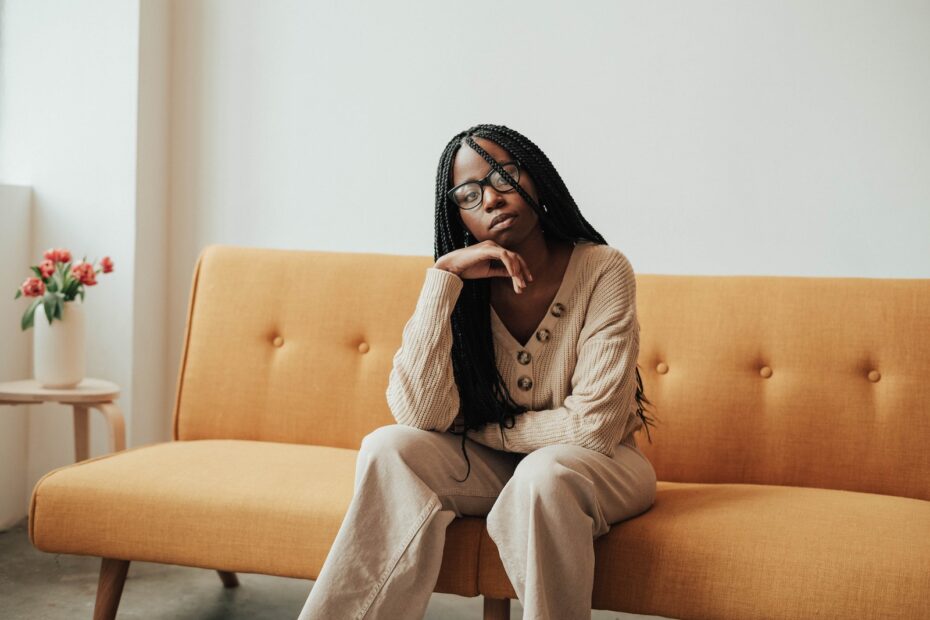Pensive young African American female in casual outfit with eyeglasses looking away while sitting on couch with hand at chin near table with flowers in vase in bright house